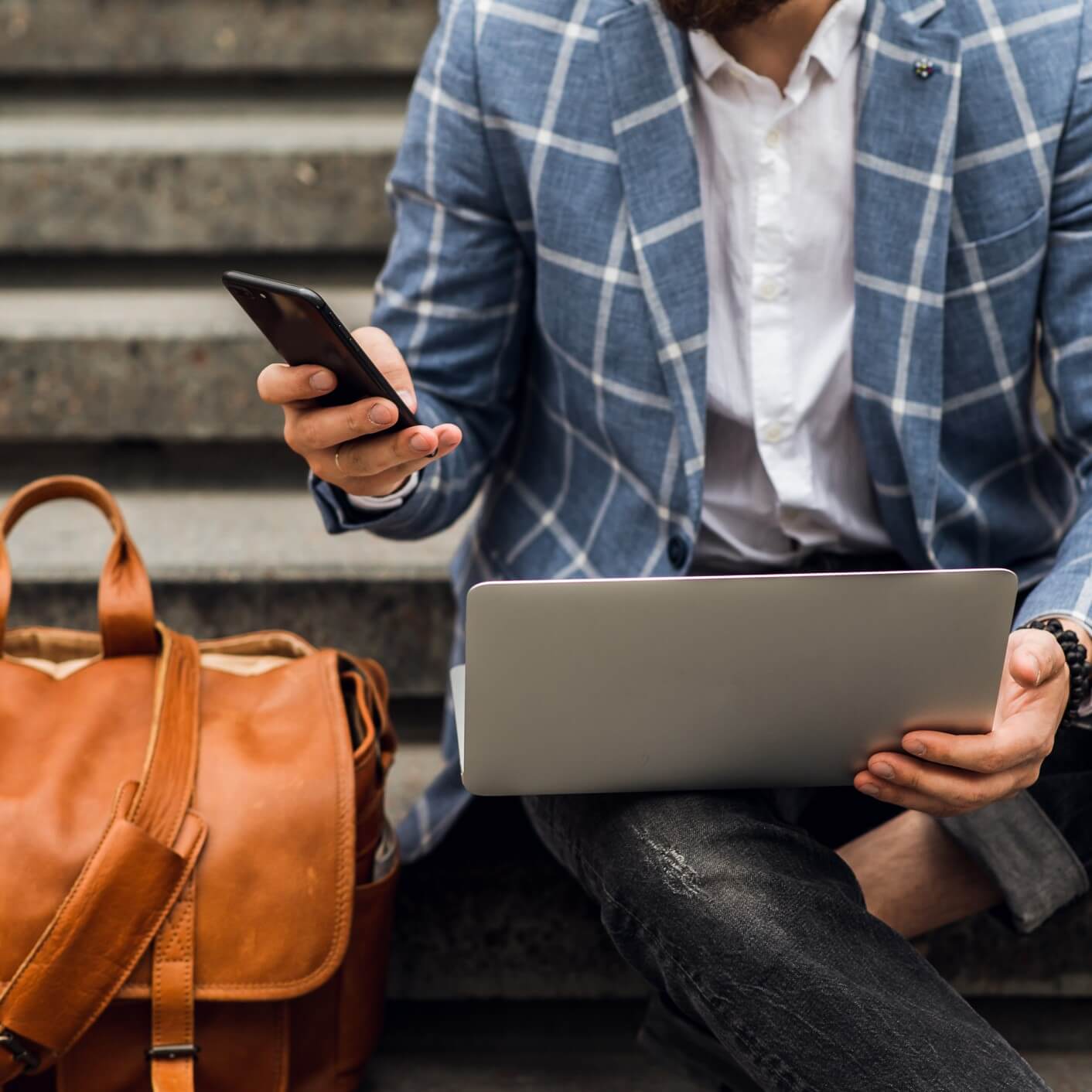 A person sits on stairs holding an open laptop in one hand and a cell phone in the other